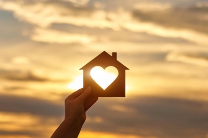 Hand holding up house-shaped cut-out with heart in the middle with sunset in background